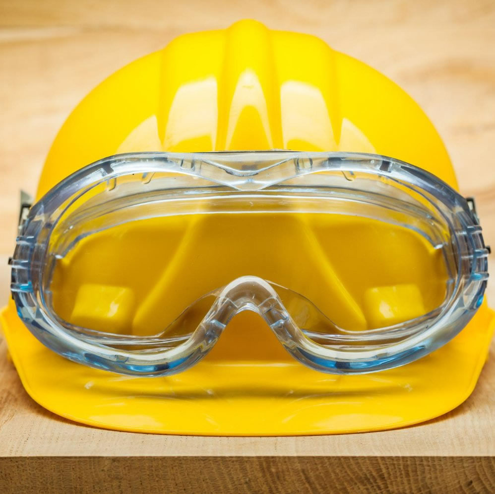 Capacete amarelo com óculos de proteção azulado por cima, ambos sobre uma mesa de madeira.